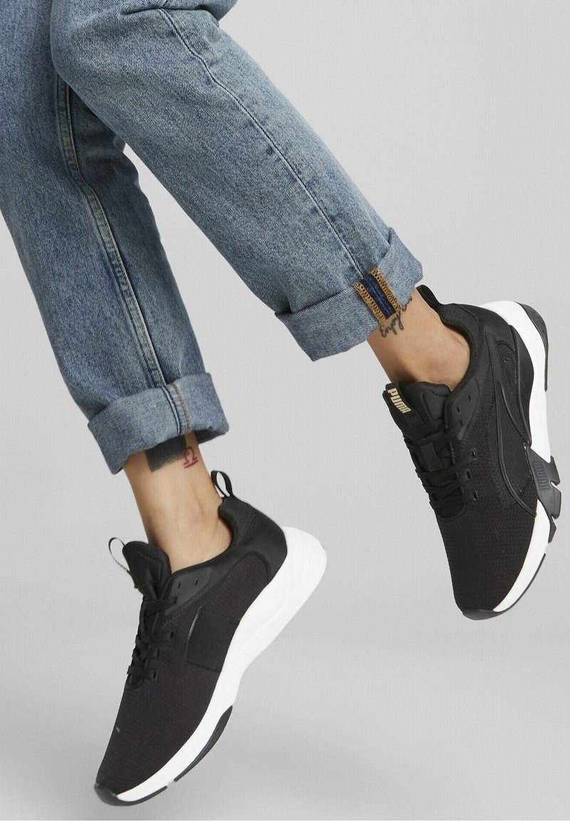 Jambes portant un jean bleu retroussé et des baskets noires Puma avec des semelles blanches sur un fond gris uni.