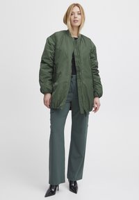 Zelená bomber bunda na zips, s elastickými manžetami a dvojicou bočných vreciek. Nosená cez čierny top a kombinovaná s tyrkysovými nohavicami s vysokým pásom.