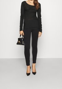 Zwarte top met lange mouwen met geplooide details, gecombineerd met nauwsluitende zwarte broek en zwarte pumps met een spits toelopende neus. Draagt een kleine zwarte handtas.