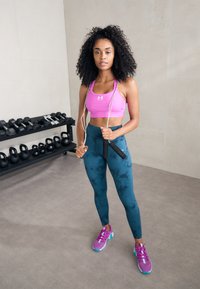 Sportliches Outfit mit einem pinken Cropped-Top, tealfarbenen, marmorierten Leggings, lila Sportschuhen und einem vor der Brust gehaltenen Springseil. Im Hintergrund sind Gewichte zu sehen.