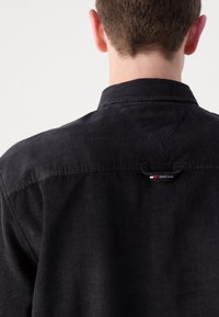 Camisa de pana negra con cuello y una pequeña etiqueta con el logo en la parte posterior, destacando la textura acanalada vertical y el color sólido.