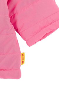 Růžový nadýchaný rukáv bundy s prošívaným vzorem, který obsahuje žlutou brandovou nášivku. Tkanina má hladkou strukturu a zaoblené okraje.