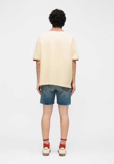 Personne aux cheveux bouclés foncés portant une chemise beige à manches courtes, un short en jean, des chaussettes à rayures orange et des baskets blanches, vue de dos.