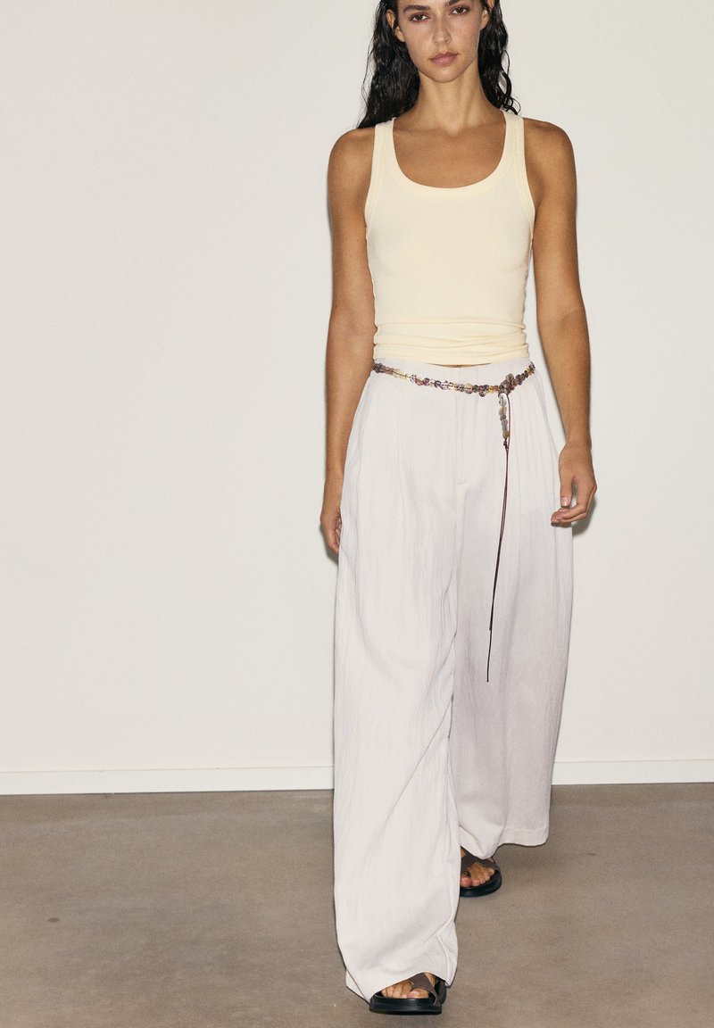 Femme portant un débardeur crème, un pantalon blanc ample avec une ceinture perlée, et des sandales noires, debout contre un mur clair uni.