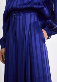 Pantalons plissés bleu vif avec des rayures verticales, en tissu brillant. Dotés d'une taille ajustée et de poches latérales.