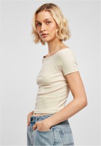 Beige ribbad t-shirt med vid halsringning och korta ärmar, figurnära silhuett, ihop med ljusblå denimjeans. Slät tygstruktur.