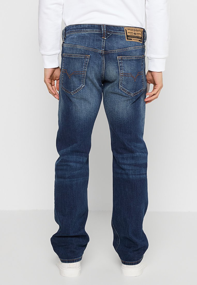 Jeans en denim bleu foncé avec une coupe droite, poches arrière visibles et une étiquette en cuir à logo sur la taille. Détails de décoloration classiques.