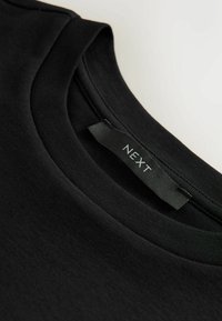 Mustast puuvillast T-särk ümmarguse kaelusega, millel on sile tekstuur ja kaela lähedal olevatel nööpidel silt "NEXT".