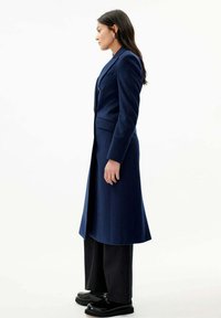 Marinblå lång kappa i en strukturerad design, med en enkel knappstängning, skräddarsydd passform och sidfickor, parat med svarta vida byxor.