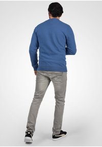 Niebieski sweter z długim rękawem i okrągłym dekoltem, noszony z jasnoszarymi dżinsami i czarnymi trampkami, pokazany od tyłu na białym gradientowym tle.