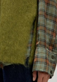 Olijfgroene donzige gebreide trui over een geruit shirt in gedempte tinten van groen, oranje en blauw, met twee knopen op de mouw.