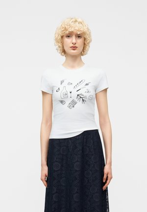 Frau mit lockigem blondem Haar trägt ein weißes T-Shirt mit schwarzen Lebensmitteldarstellungen und einen dunklen, gemusterten Rock vor einem schlichten Hintergrund.