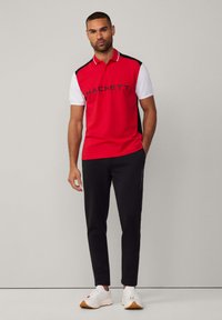Rotes Poloshirt mit kurzen weißen Ärmeln und schwarzen Akzenten, mit "HACKETT"-Logo. Schwarze Sporthose und weiße Turnschuhe runden das Outfit ab.