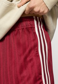 Rdeče športne kratke hlače z navpičnim vzorcem črt in belimi stranskimi črtami, z elastičnim pasom in mehko teksturo.