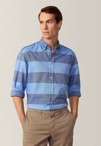 Camisa de botones a rayas azul claro y azul oscuro con mangas remangadas, hecha de tejido entrelazado, combinada con pantalones caqui.
