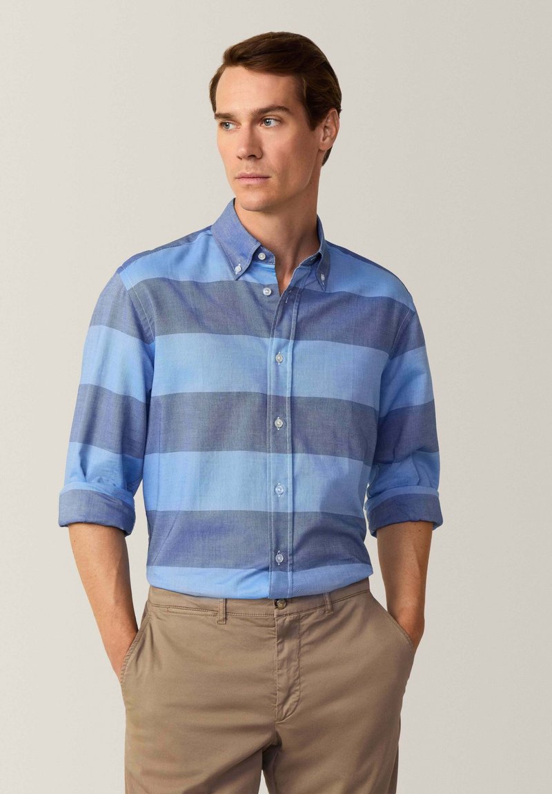 Camisa de botones a rayas azul claro y azul oscuro con mangas remangadas, hecha de tejido entrelazado, combinada con pantalones caqui.