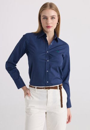 Femme aux cheveux blonds lisses portant une chemise bleu marine à boutons et un pantalon blanc avec une ceinture tressée marron, debout devant un fond uni.