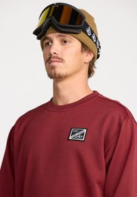 Sweatshirt bordeaux avec un col ras du cou et un patch logo Volcom. L'individu porte un bonnet en tricot marron et des lunettes de ski avec des verres miroités.