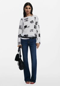 Cămașă albă cu mânecă lungă, decorată cu modele grafice negre, asociată cu blugi evazați albaștri. Modelul ține o poșetă neagră și poartă sandale cu toc roșii.