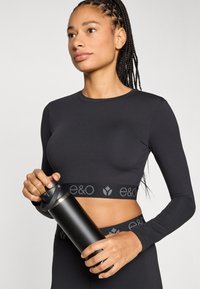 Μαύρο κορμάκι με μακριά μανίκια και λογότυπο στη ζώνη, εφαρμοστή σχεδίαση και λεία υφή. Το άτομο κρατά ένα μαύρο μονωμένο μπουκάλι νερού.