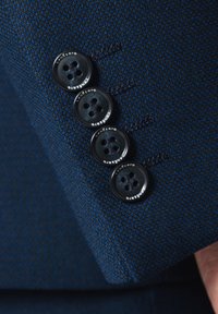 Navy teksturowana tkanina z czterema czarnymi guzikami na mankiecie, każdy z czterema otworami i grawerowanym logo. Zbliżenie detaliczne na szycie.