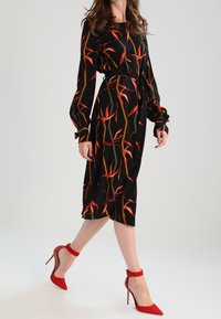 Schwarzes Midi-Kleid mit langen Ärmeln, versehen mit einem Blumenmuster in Rot und Orange. Kombiniert mit roten hohen Schuhen.