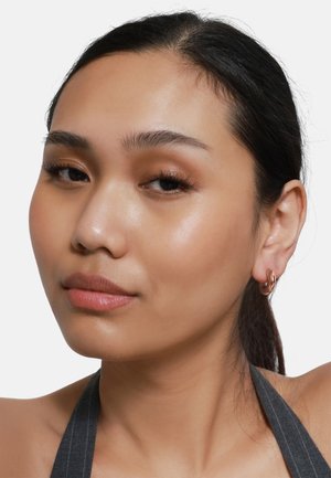 Nahaufnahme eines Gesichts mit glatter Haut, dunklem Haar und einem roségoldenen Creolenohrring. Der Hintergrund ist schlicht weiß.