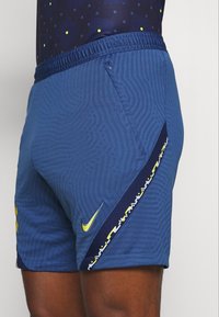 Pantaloni sportivi blu con un motivo testurizzato, dotati di un logo Nike giallo e una striscia accentuata blu navy con elementi di design geometrici.