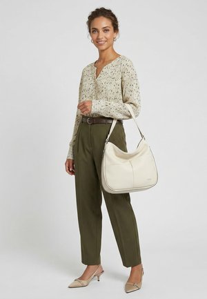 Frau steht, trägt eine beige gemusterte Bluse, olivgrüne Hose, beige High Heels und hält eine große weiße Umhängetasche.