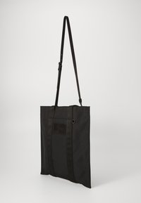 Černá látková taška s dlouhým nastavitelným popruhem, obdélníkového tvaru, se dvěma vrchními držadly a malou nášivkou na přední straně. Hladký povrch.