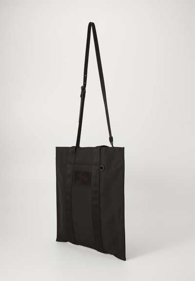 Black fabric tote bag with long adjustable strap, rectangular shape, two top handles, and a small patch on the front. Smooth texture.