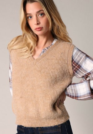 Gilet en tricot beige avec un col en V, fabriqué en fil texturé, superposé sur une chemise à carreaux bleue et blanche, présentant une coupe décontractée et un motif côtelé.