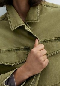Ruka vytahující přední část olivově zelené bundy s klopovými kapsami a viditelným švem.