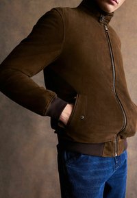 Ruskea mokkanahkainen bomber-takki, jossa korkea kaulus, vetoketjullinen etuosa, resorit hihansuissa ja sivutaskut. Yhdistetty sinisiin farkkuihin.