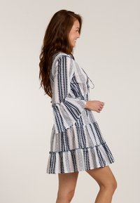 Weißes Kleid mit blauen geometrischen Mustern, weit ausgestellten Ärmeln und einem Rüsche-Saum. Mit einem Kordelzug am Ausschnitt zur Anpassung gestaltet.