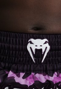 Σκούρο δέρμα στην κοιλιά πάνω από μαύρη ελαστική μέση με λευκό στυλιζαρισμένο λογότυπο φιδιού ή πλάσματος με δόντια, κάτω από μωβ και μαύρα καμουφλάζ σορτς.