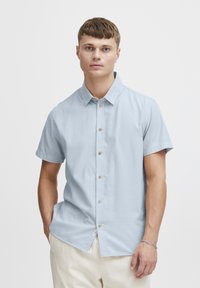 Lichtblauw kortverzekering shirt van gladde stof, met een knoopsluiting aan de voorkant, lichtbruine knopen en een klein zijlabel detail.