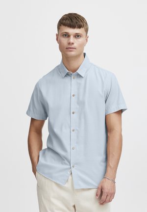 Chemise à manches courtes bleu clair en tissu lisse, dotée d'un devant à boutons, de boutons marron clair et d'un petit détail d'étiquette sur le côté.