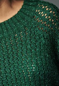 Pull en tricot vert avec un tissage lâche et ouvert, orné de sequins brillants, dotée d'un col côtelé et d'une texture légèrement transparente.