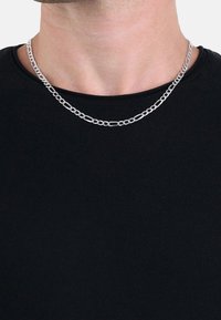 Gros plan sur une personne portant un collier chaîne argenté avec des maillons ovales et ronds entrelacés sur une chemise noire.