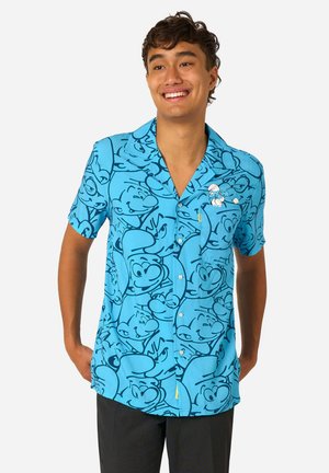 Jonge man die lacht en een blauw shirt met korte mouwen draagt met meerdere cartoon Smurfen gezichten en één Smurf geborduurd op het borstzakje.