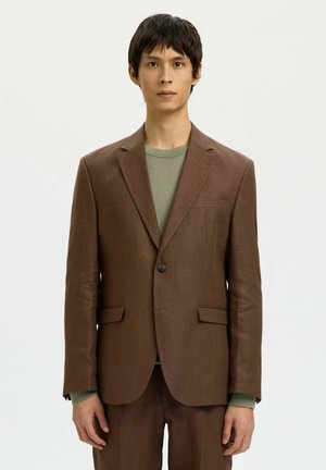 Homme portant une veste de costume marron à un bouton et un pantalon assorti, avec un haut vert à col rond, debout devant un fond uni.