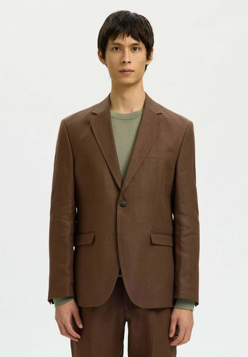 Homme portant une veste de costume marron à un bouton et un pantalon assorti, avec un haut vert à col rond, debout devant un fond uni.