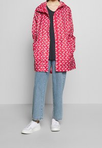 Czerwony płaszcz przeciwdeszczowy z powtarzającym się wzorem psów, luźny krój i kieszenie, w połączeniu z czarną bluzką i jasnoniebieskimi dżinsami. Całość uzupełniają białe sneakersy.