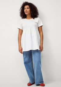 Hellgraues, kurzärmeliges Oberteil mit gerafftem Bund, kombiniert mit hellblauen Schlagjeans. Rote Schuhe runden das Outfit ab. Flache Textur.