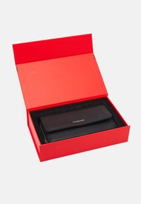 Czarny skórzany portfel z złotym logo w czerwonym pudełku; charakteryzuje się eleganckim wzornictwem i zamknięciem na zamek, z miękkim czarnym wnętrzem.