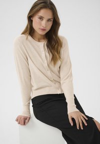 Cárdigan beige de punto con escote redondo, con botones en la parte delantera. Mangas largas con puños acanalados, combinado con una falda negra.
