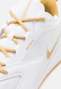 Hvit fritidssko med gullakenter, med en strukturert mesh-tunge, vevde gullsnøringer, og "JUST DO IT." i gull på siden.