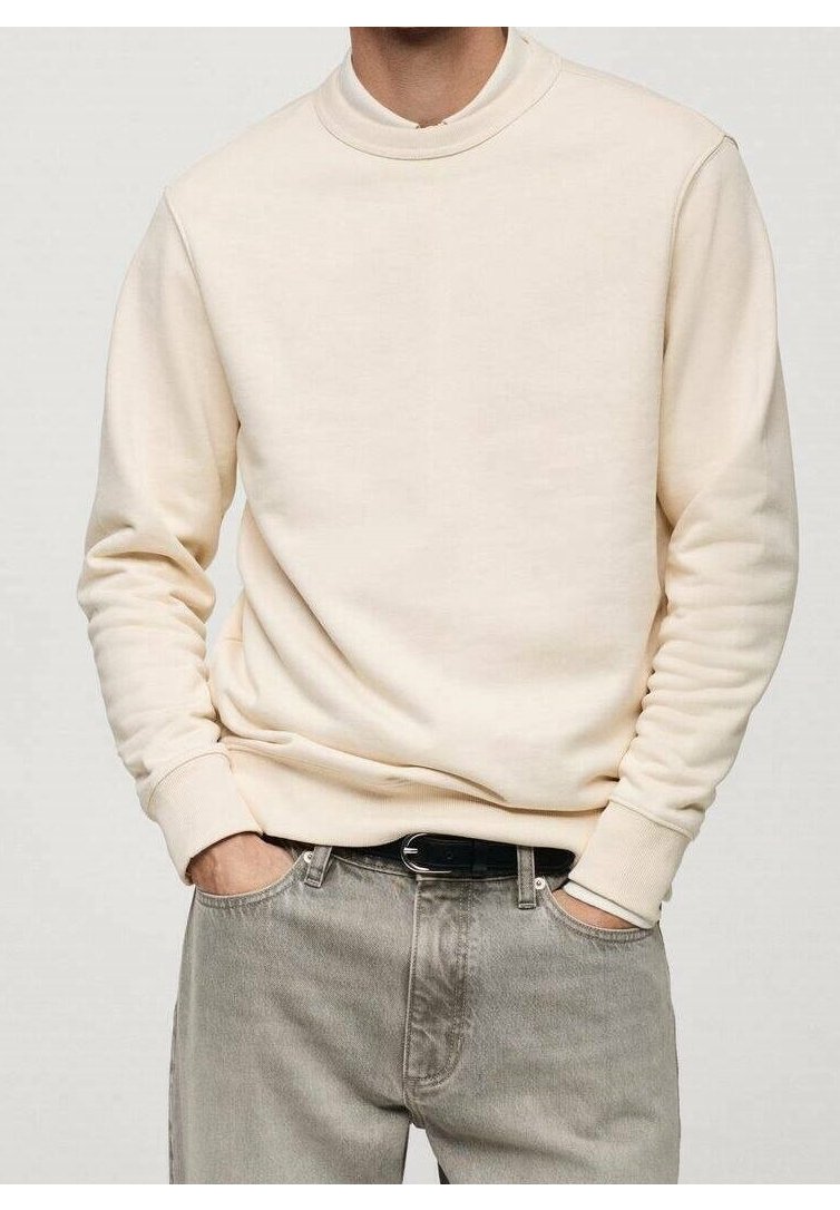 Homme portant un sweat-shirt couleur crème et un jean gris, les mains dans les poches, debout devant un fond uni.