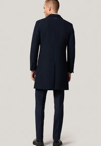 Granatowy wełniany płaszcz o strukturalnym designie, z pojedynczym rozcięciem z tyłu i zapięciem na guziki przy mankietach. Noszony na dopasowanych spodniach i czarnych butach.